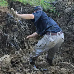 A plumber in a navy shirt and cap working a digging bar through tree roots in a water-line dig.