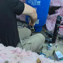 A plumber kneeling to fit piping at a blue well pressure tank.
