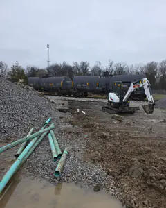 A broken 4-inch line being repaired beside a rail line, a mini excavator and green pipe in the mud.